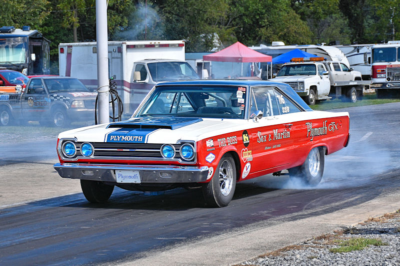A vintage Plymouth GTX muscle car performing a burnout on a drag strip, with smoke rising from the rear tires and a clear blue sky in the background.