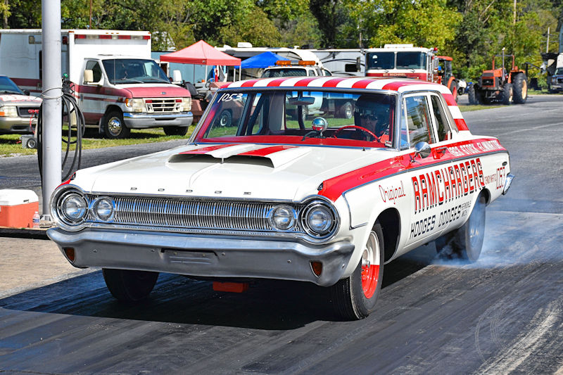 A vintage Dodge racing car with a red and white color scheme, seen on a drag strip, with smoke coming from the rear tires.