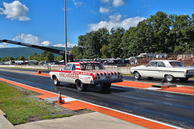 A drag racing scene featuring a vintage car with red and white stripes on the track, while another classic car is positioned nearby. The background shows spectators and trees under a clear blue sky.