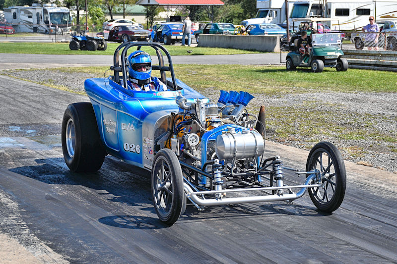 A blue drag racing car on a track, featuring a driver in a helmet and racing suit, with a detailed engine visible in the front.