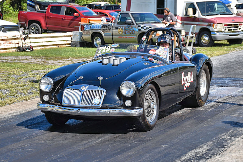 A vintage black MG car racing down a drag strip, with a driver in a helmet and racing gear. The car features racing decals and has an open cockpit design.