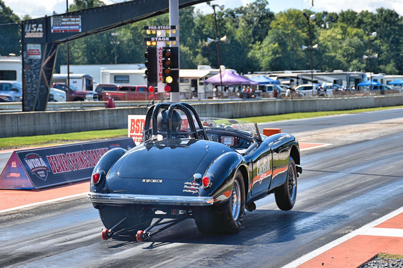 A black vintage sports car racing down a drag strip with a drag racing tree illuminated in the background.