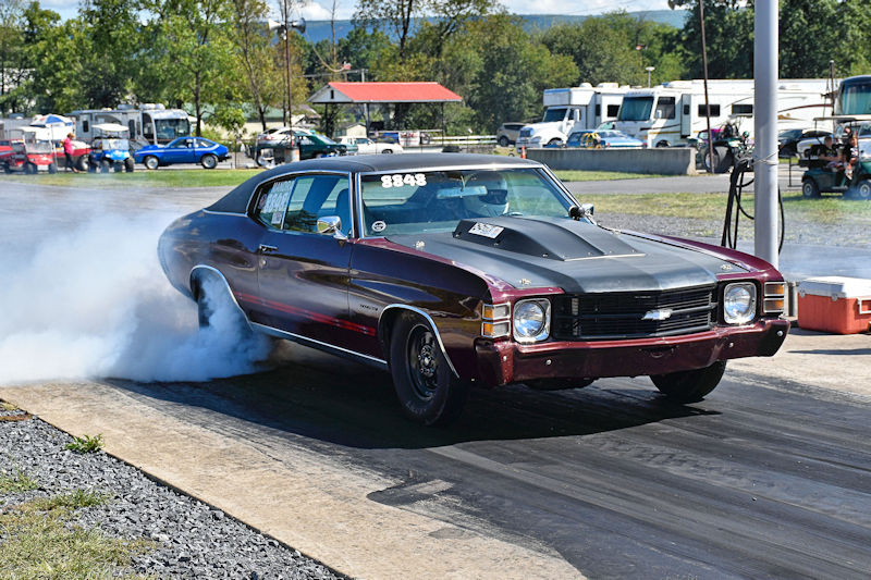 A classic Chevrolet muscle car performing a burnout on a drag strip, with smoke billowing from the rear tires.