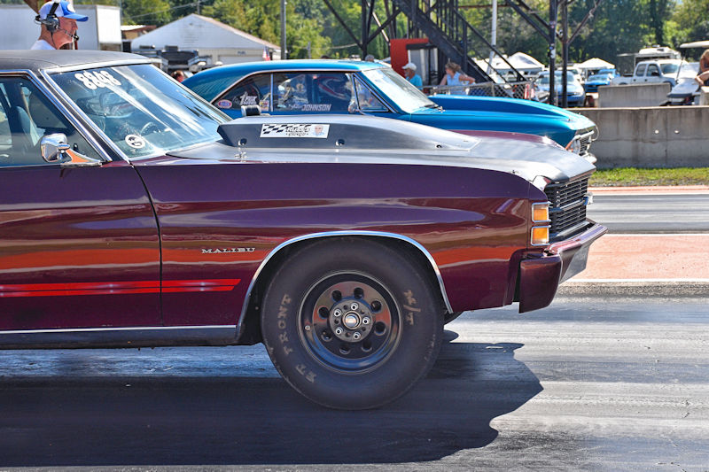 Side profile of a red and black vintage car, with a large rear tire, positioned on a drag racing strip with another car in the background.