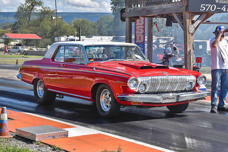 A vintage red and white muscle car with a hood scoop is positioned on a drag racing strip, showcasing its sleek design under clear skies. In the background, a spectator is taking photos.