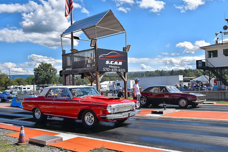 A red classic car accelerating down a drag strip, with a dark-colored car in the background, a timing tower in the foreground, and an American flag flying above.