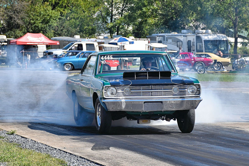 A green and black classic muscle car performing a burnout on a drag strip, surrounded by a crowd of spectators and parked vehicles in the background.
