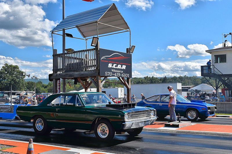 A green classic car and a blue car lined up on a drag racing strip, with spectators and a timing tower in the background under a partly cloudy sky.
