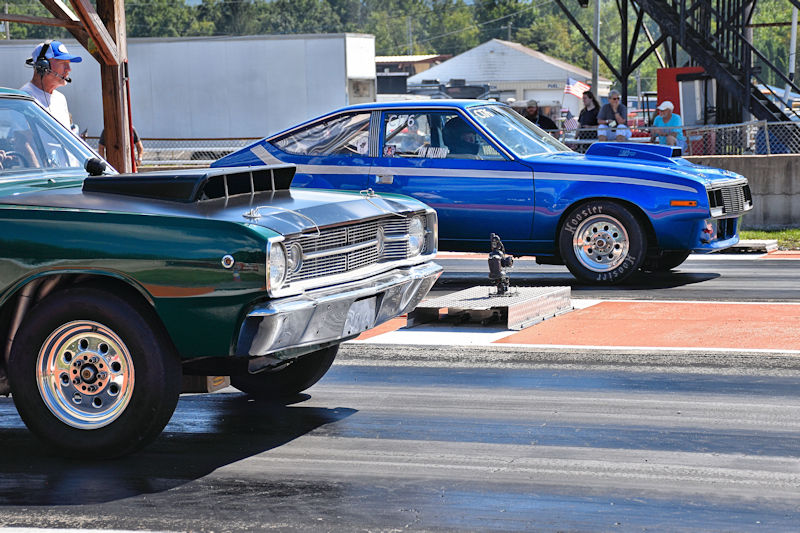 A green classic car and a blue racing car lined up at a drag strip, with spectators in the background.