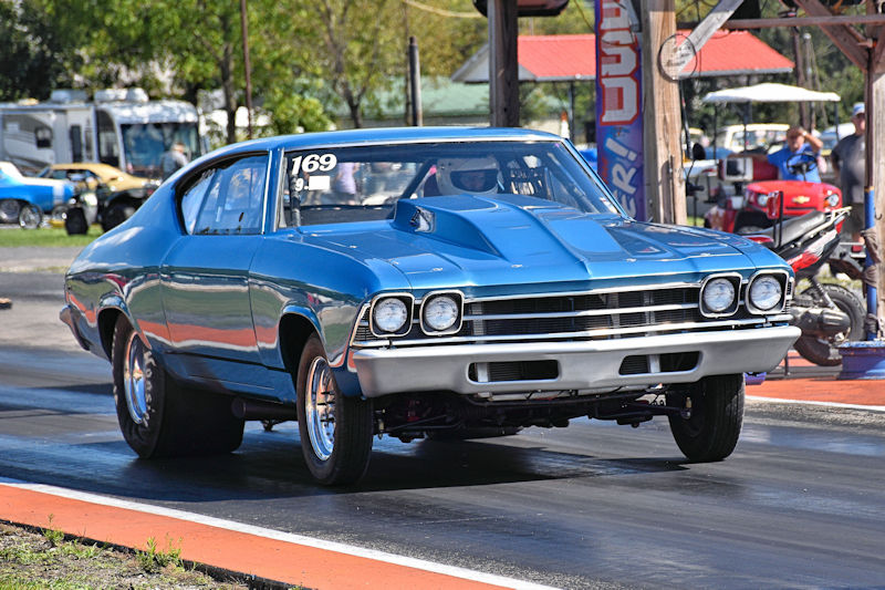 A blue vintage car with a racing number prominently displayed on the windshield, speeding down a drag strip.