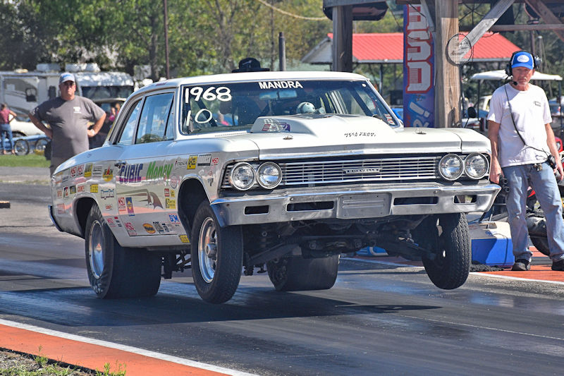 A vintage 1968 Chevrolet sedan performing a wheelie on a drag strip, with various sponsorship decals visible on its sides. In the background, spectators can be seen watching the event.