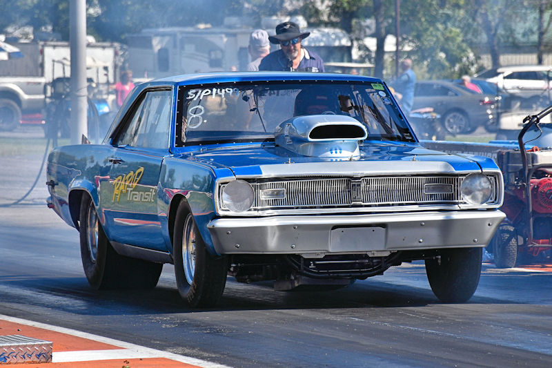 A blue vintage drag racing car with a hood scoop, accelerating on a racetrack, with smoke rising from the tires and spectators in the background.