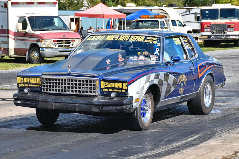 A custom drag racing car with a blue and white design featuring 'Moe's Auto Repair' signage parked on a racetrack, surrounded by various trailers and support vehicles.