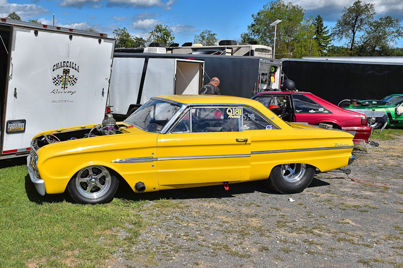 A classic yellow racing car parked on a grassy area with a trailer in the background. The car has a supercharged engine and large rear tires, showcasing a custom racing design.