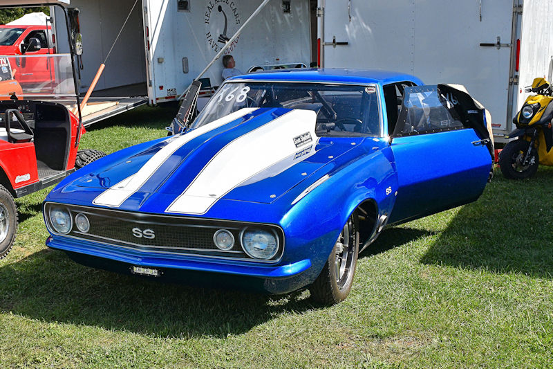 A blue Chevrolet Camaro SS with white racing stripes, parked with an open door, on green grass at an outdoor event.