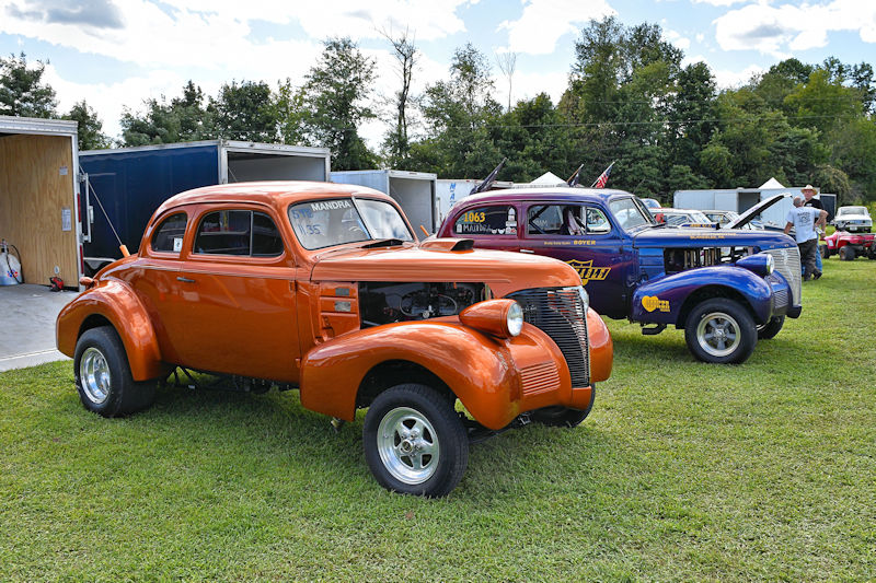 Two classic modified cars parked on green grass at an outdoor event, featuring an orange coupe in the foreground and a blue coupe behind it, with a cloudy sky in the background.