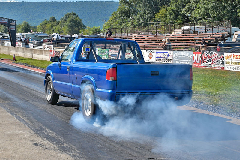 A blue pickup truck performing a burnout on a drag racing track, producing smoke from its tires.