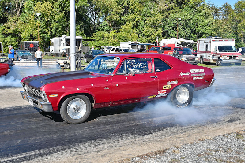 A red Chevrolet Nova drag racing car with smoke billowing from the tires, speeding down a drag strip during a race event.