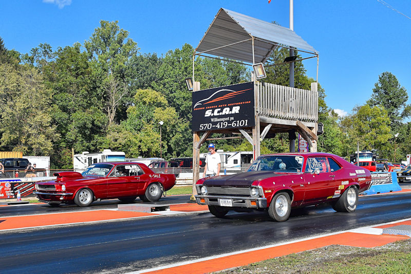 Two vintage drag racing cars lined up on a racetrack, with a timing tower in the background and spectators nearby.