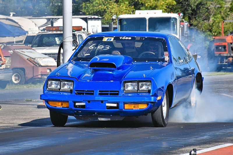 A blue drag racing car with a hood scoop accelerates down a racetrack, leaving a trail of smoke behind.