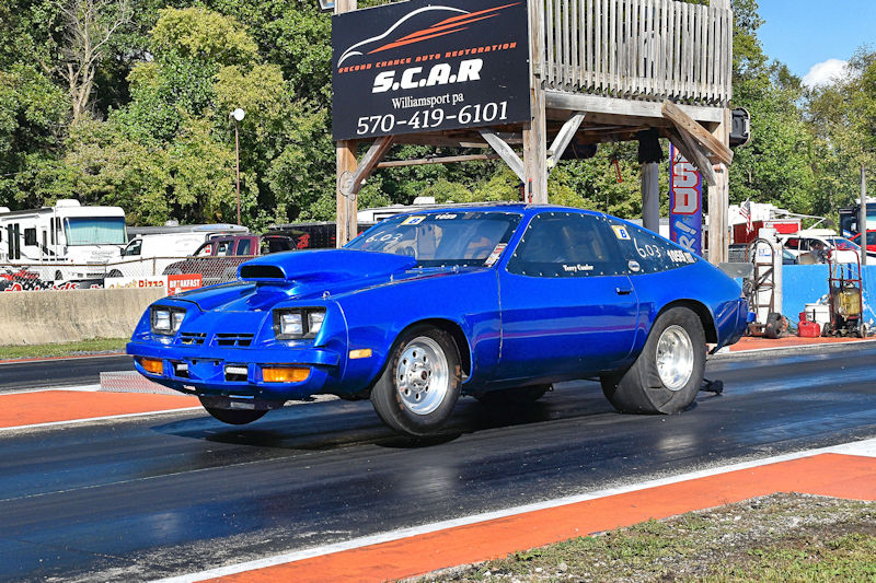 A bright blue drag racing car launching from a starting line at a racetrack, with a wooden structure and various vehicles in the background.