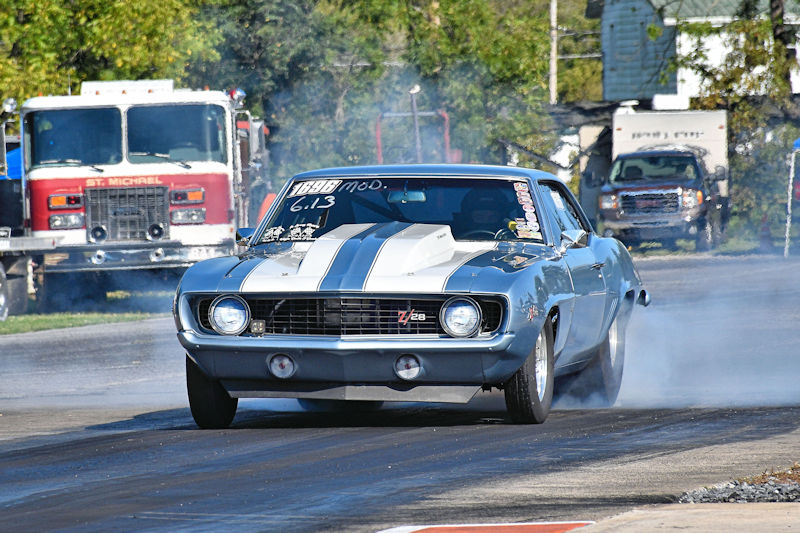 A classic gray Chevrolet Camaro Z28 with white stripes performing a burnout on a drag racing track, surrounded by trees and vehicles in the background.
