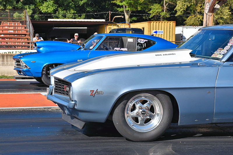 Two classic muscle cars lined up on a drag strip, one silver with a white hood labeled 'Z/28' and the other blue, ready for a race.