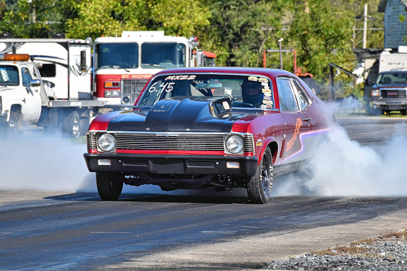 A classic drag racing car executing a burnout on a racetrack, surrounded by vehicles in the background.