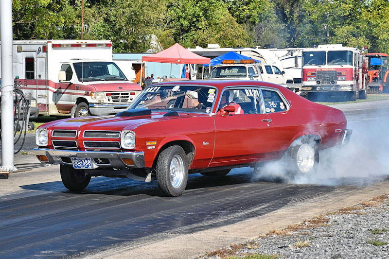 A classic red muscle car accelerating on a drag strip, producing tire smoke, with support vehicles and trailers in the background.