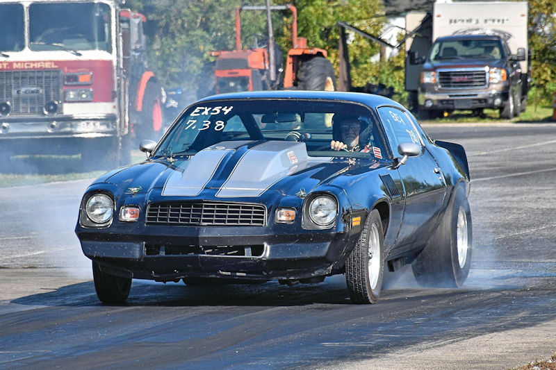A classic black race car with a silver stripe is performing a burnout on a drag strip. Smoke is billowing from the rear tires as the car accelerates, showcasing its racing capabilities.