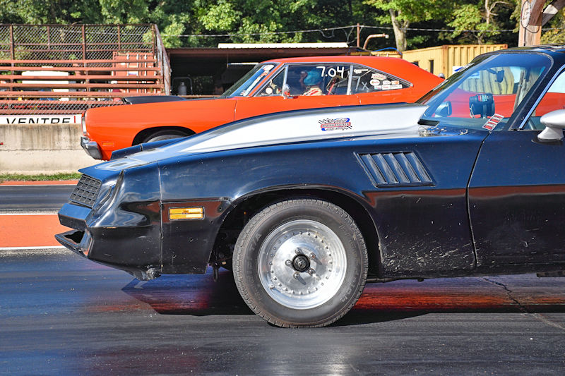 Side view of two race cars lined up at a drag strip, featuring a black car with a silver hood and an orange car in the background.