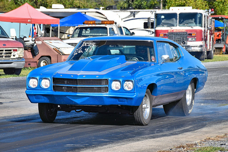 A blue classic car launching off the starting line at a drag racing event, with smoke from the rear tires and various vehicles and tents in the background.