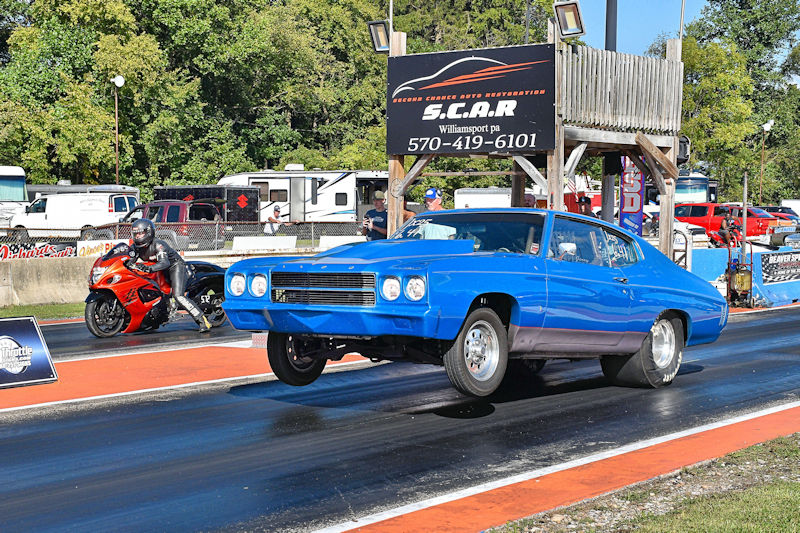 A blue classic muscle car performing a wheelie on a drag strip, with a motorcycle racer in the background preparing for a race.