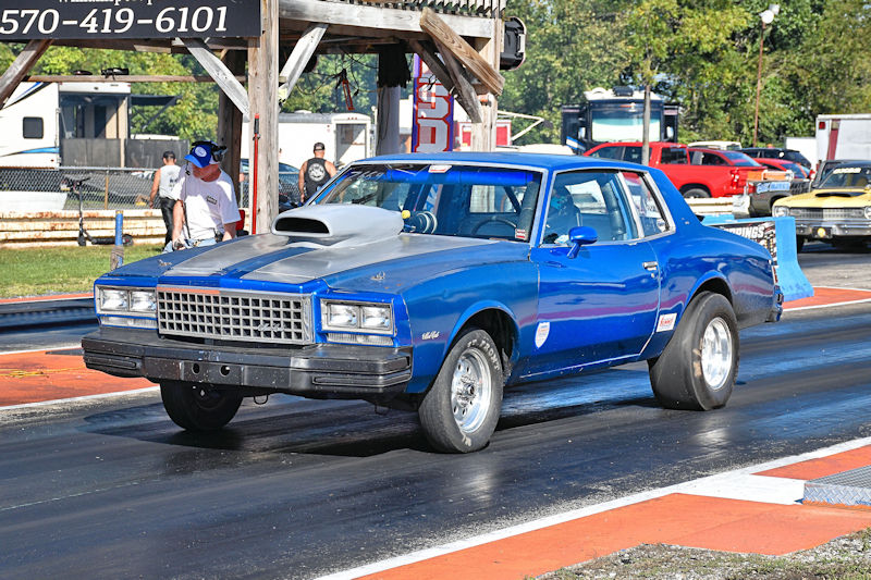A blue racing car with a prominent hood scoop on a drag strip, showcasing high-performance features, with spectators and other vehicles in the background.