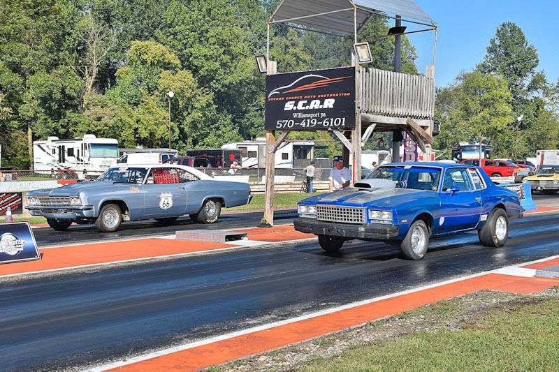 Two classic cars lined up on a drag strip, one gray and one blue, with a timing tower in the background.