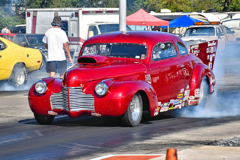 A bright red vintage drag racing car performing a burnout on a racetrack, with smoke billowing from the tires. In the background, other cars and tents are visible, along with a person walking in the foreground.