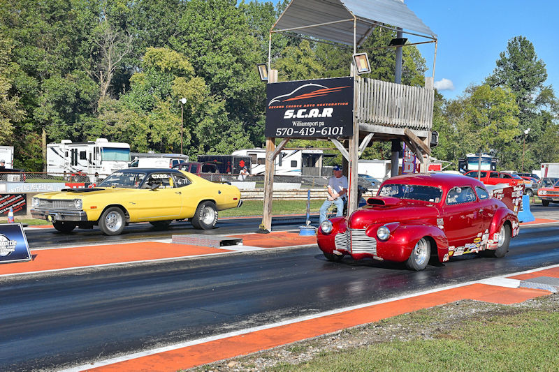 Two cars lined up on a drag racing track, with a yellow car on the left and a red car on the right, while spectators and a race official are nearby.