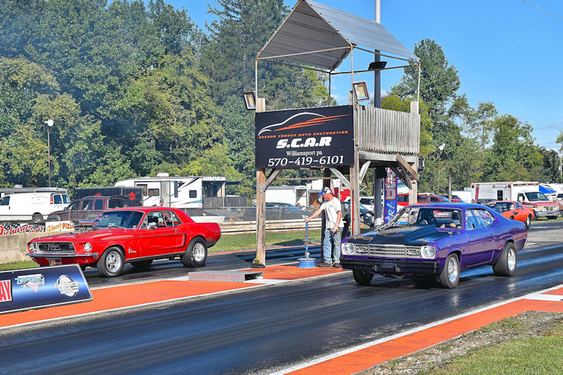 A red classic Mustang and a purple race car lined up on a drag strip, with a track official preparing the start. A sign displaying 'S.C.A.R.' is visible in the background, alongside trees and parked vehicles.