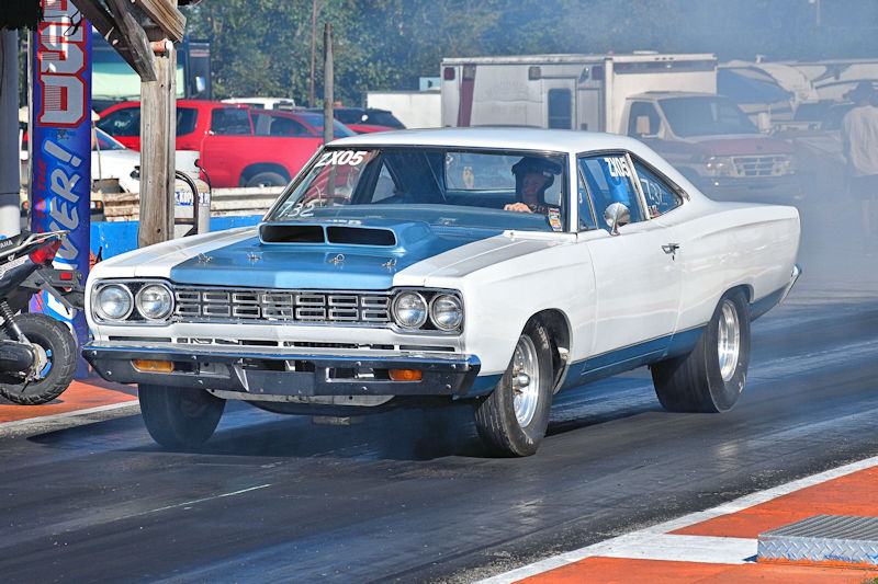 A classic white and blue muscle car accelerating down a drag racing track, with smoke trailing behind the rear tires.