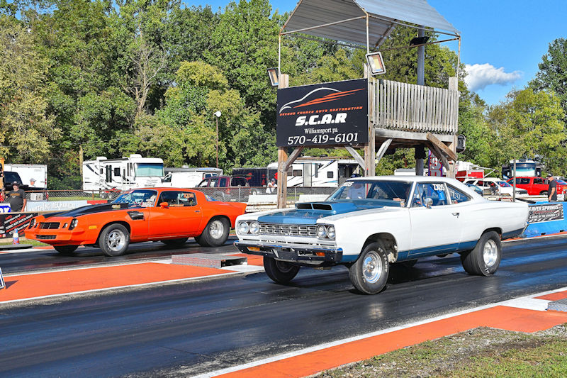 Two classic muscle cars lined up on a drag racing track, one in orange and one in white, with a timing tower and green trees in the background.