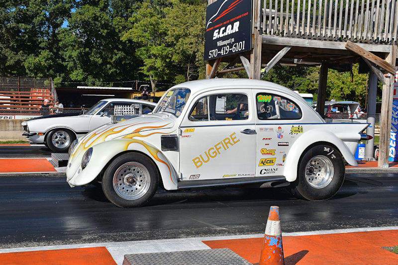 A vintage white Volkswagen Beetle race car with flame graphics, featuring the name 'BUGFIRE,' on a drag racing track with a grey muscle car visible in the background.