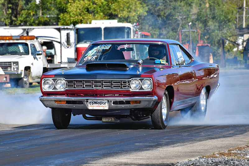 A vintage red muscle car performs a dramatic burnout on a racetrack, emitting smoke from its rear tires. The car features a hood scoop and a sleek design, showcasing its racing modifications.