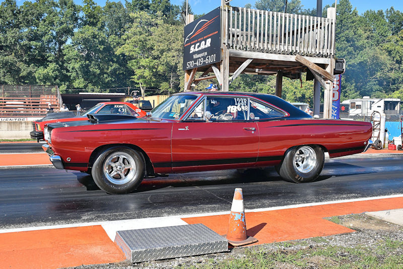 A classic red muscle car preparing to race on a drag strip, with a racing starting line and a timing tower in the background.
