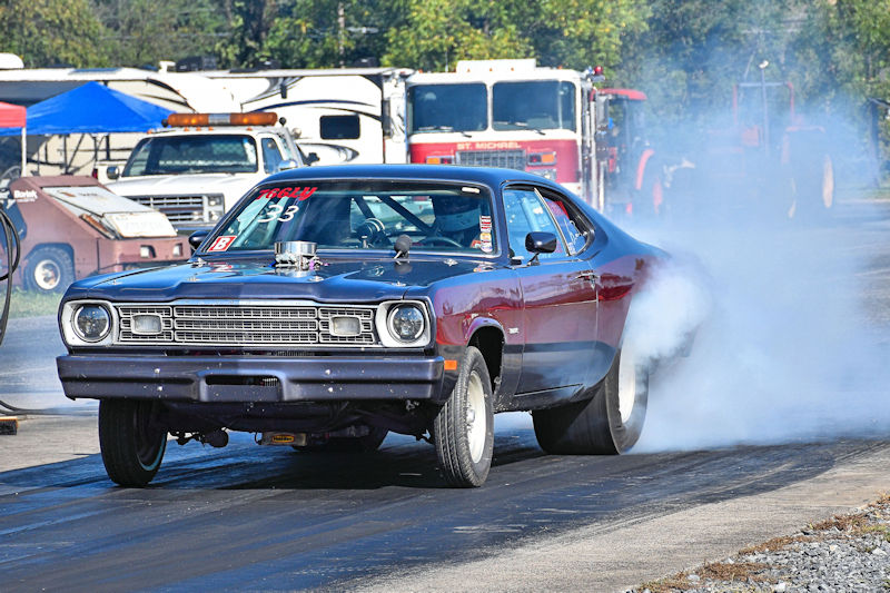 A classic muscle car with a dark blue and maroon exterior is performing a burnout on a drag strip, with smoke emanating from the rear tires.