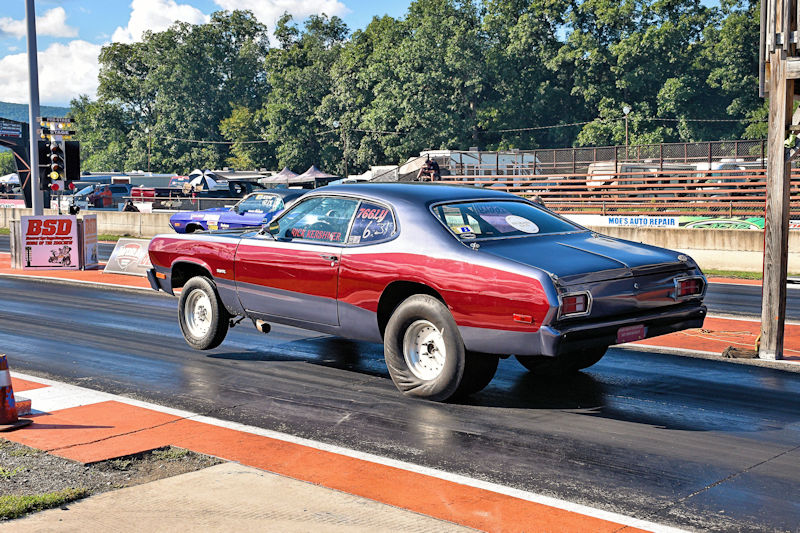 A classic drag racing car with a red and silver color scheme accelerating down a drag strip, with trees and a racetrack in the background.