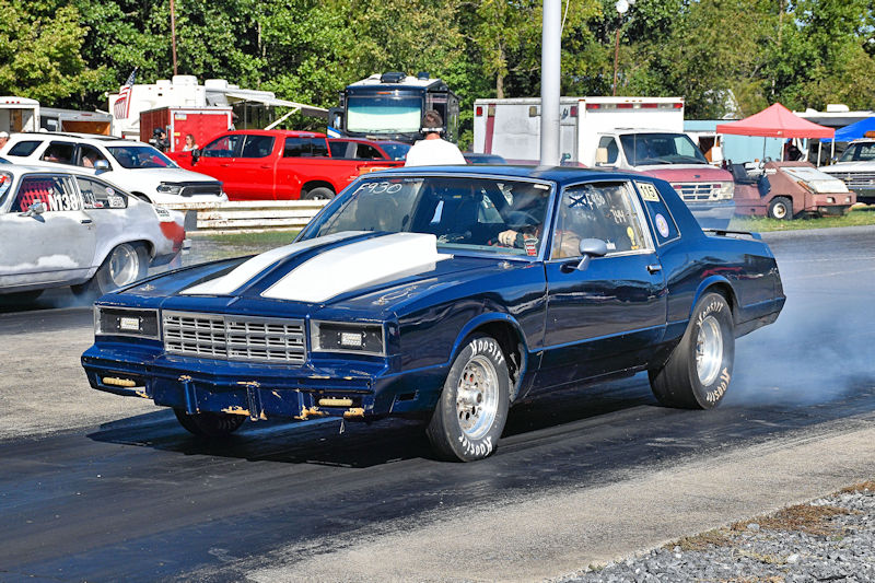 A blue drag racing car with a white hood stripe racing down a track, creating smoke from its tires, with spectators and other vehicles in the background.