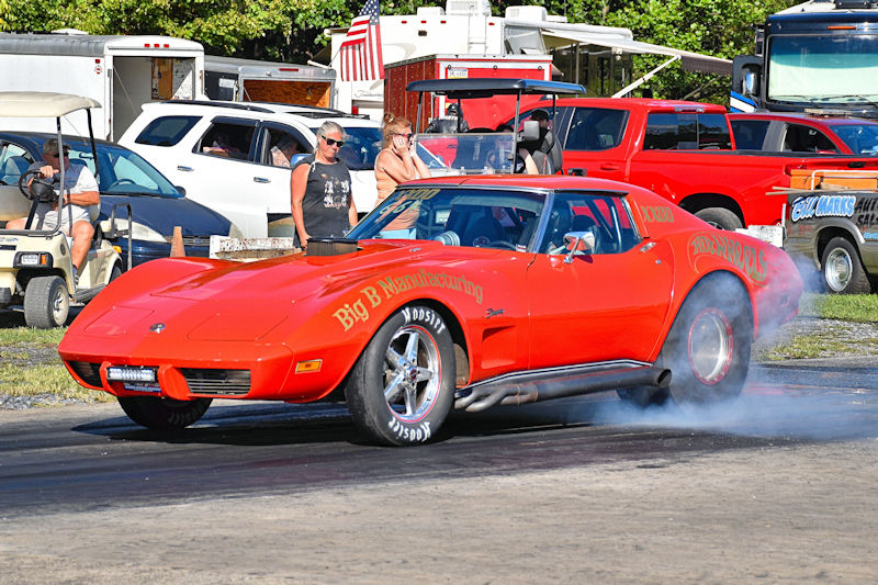 A bright orange vintage Corvette racing car emitting smoke from its tires at a drag strip, with spectators in the background.