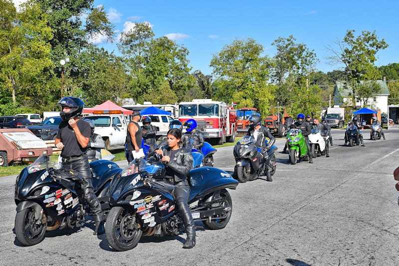 A group of motorcycle riders in riding gear preparing to race on a sunny day, with various motorcycles lined up in the background.