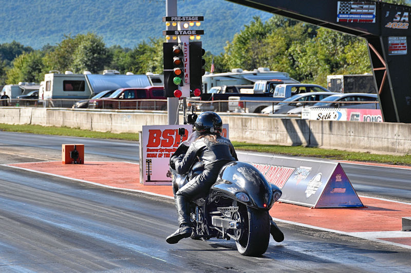 A motorcycle racer in a black leather suit prepares to launch off the starting line at a drag strip, with timing lights in the background and spectators' vehicles parked nearby.