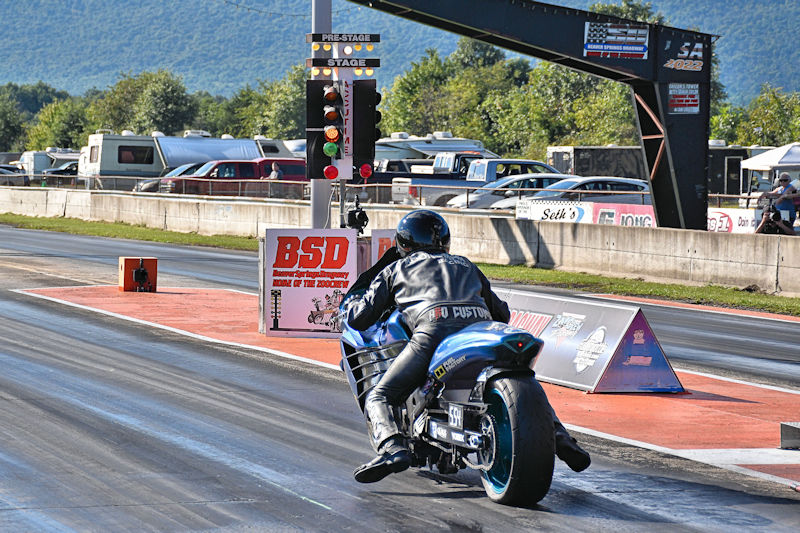 A motorcycle racer wearing a black leather suit prepares to race down a drag strip, with green traffic lights visible in the background and spectators in the distance.